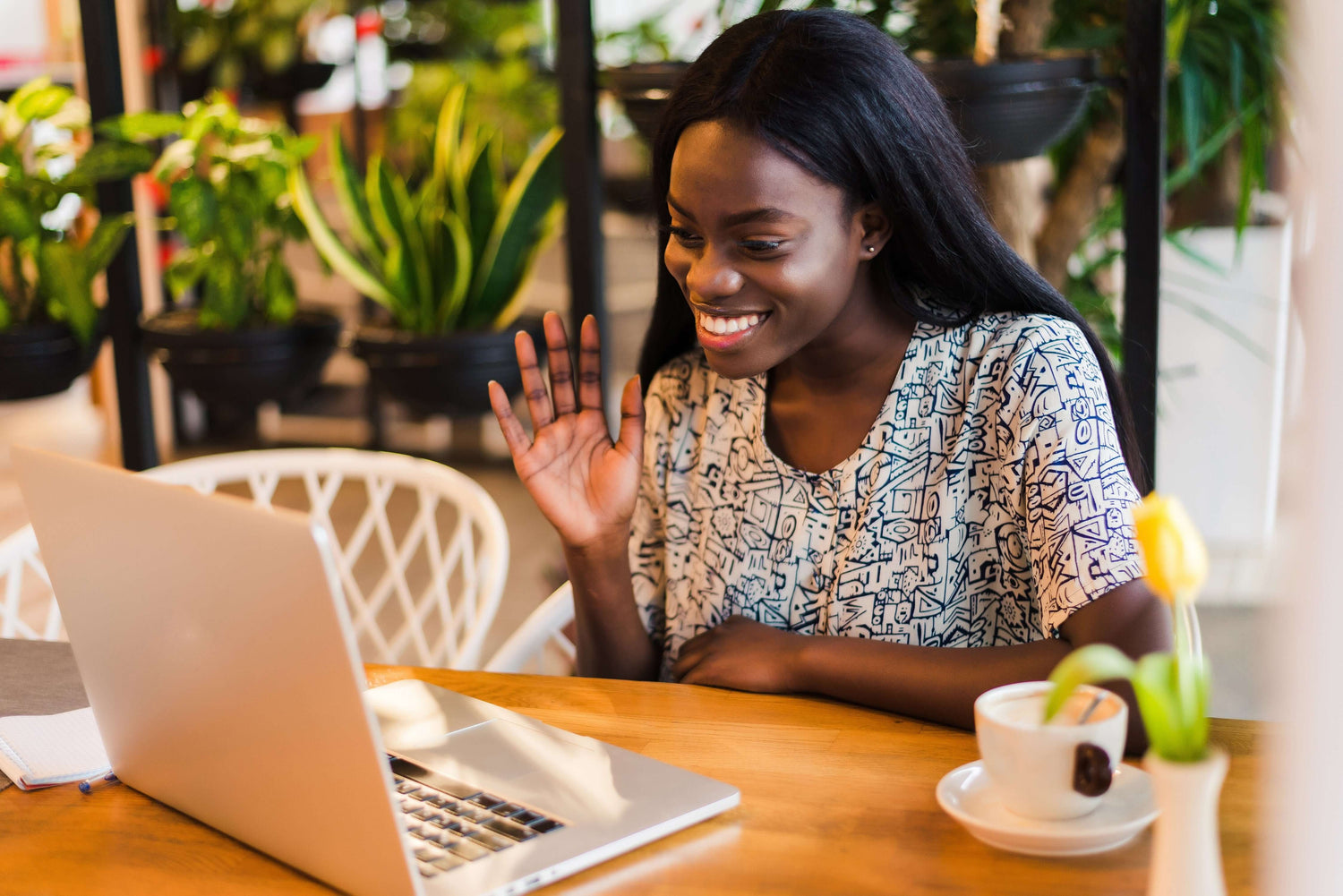 Woman smiling and waving while attending English speaking lessons online at a cozy café.
